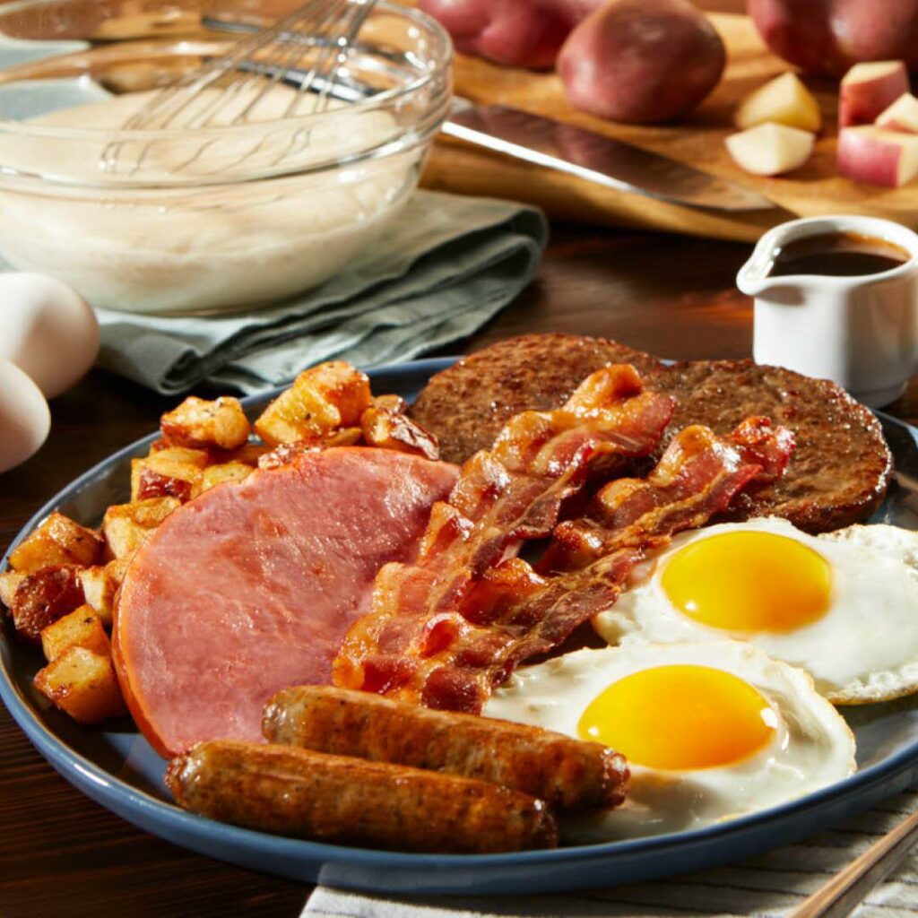 Breakfast platter that includes bacon, sausage, ham, sausage links, and eggs with food in the background