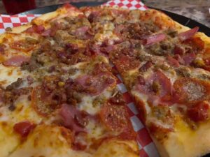 Pictured here is a mouth watering view of The Meats Pizza-with chuncks of Sausage, Beef, Bacon, and Ham along with slices of Pepperoni on a bed of cheese, pizza sauce, and thin crust.