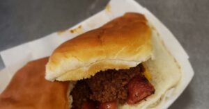 A paper tray holding a mouth watering sandwich on a hotdog bun stuffed with a crsipy hotdog smothered in chili sauce and a slice of cheese underneath.