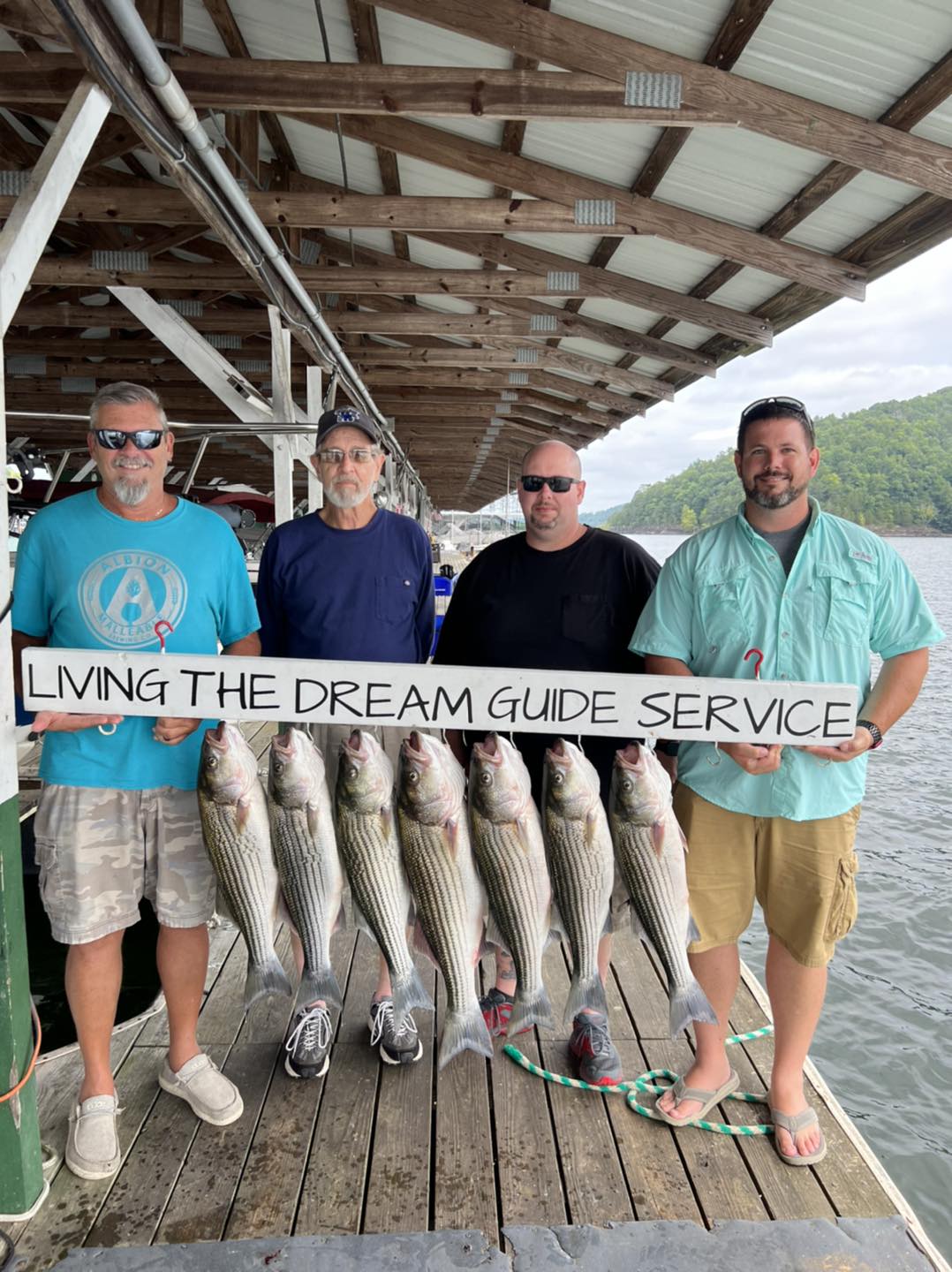 Four men holding onto a board that says Living the Dream Guide Service, has seven striper fish hanging below.