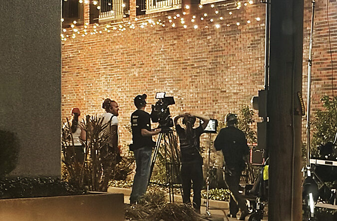 men and women on a film set with cameras and lights, standing in an alleyway between two buildings with string lights above them.